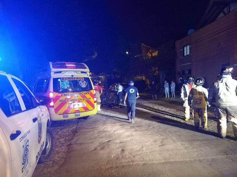 Bomberos en uniforme interactúan en la escena de un accidente, con una ambulancia visible y espectadores cerca, en una noche iluminada.