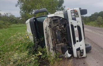 Camión volcado en la orilla de una carretera rural con cielo nublado y vegetación densa alrededor.