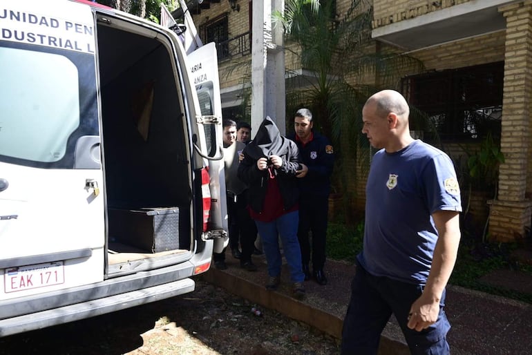 Hombre encapuchado y vestido con sudadera oscura es custodiado por agentes de seguridad cerca de un vehículo policial.