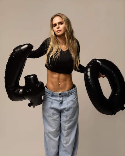Mujer con cabello largo y lacio, viste blusa negra y pantalones de denim, sostiene globos negros con el número 40.