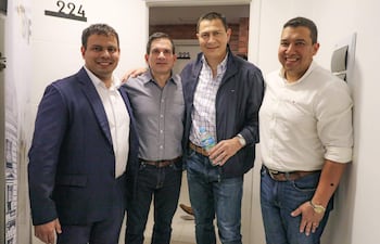 Raúl Salinas y otros tres hombres sonrientes, de pie en una oficina, vistiendo trajes oscuros y chaquetas, con una botella de agua entre ellos.