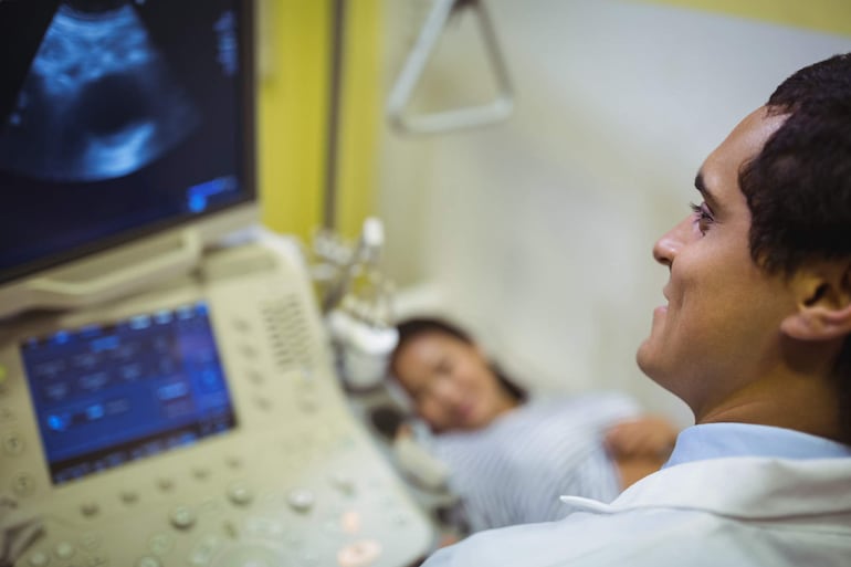 Médico en bata blanca observando pantalla de ecografía mientras mujer recostada en camilla viste camiseta de rayas.