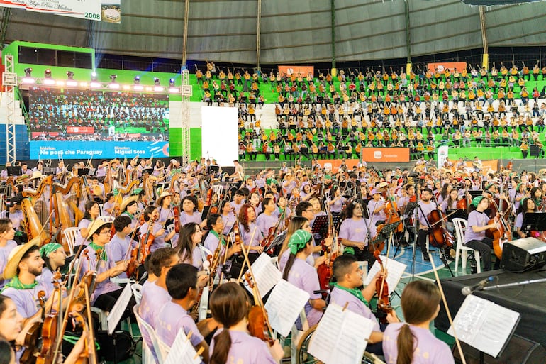 Jóvenes músicos con camisetas moradas y algunos con pañuelos verdes, interpretando en un gran escenario con un monitor visible.