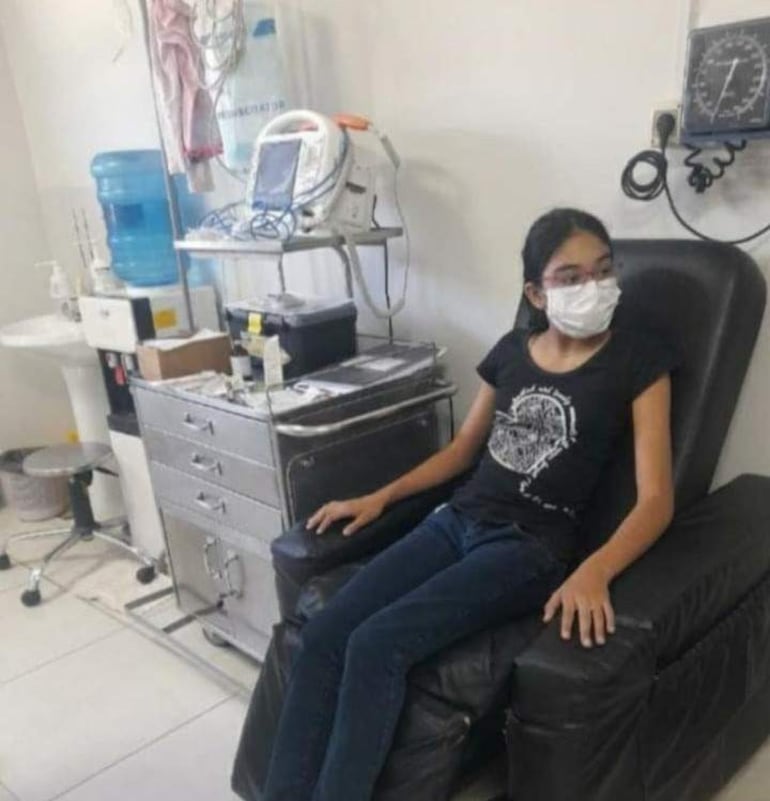 Niña de 14 años con camiseta negra y tapabocas blanco, sentada en un sillón en un consultorio médico, rodeada de equipos clínicos.