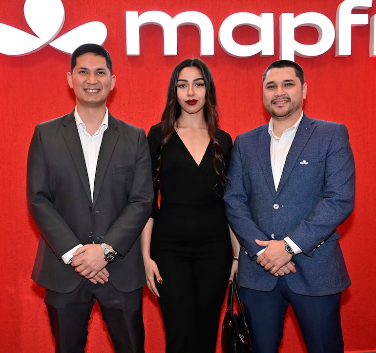 Mujer con mono negro y labios rojos, flanqueada por dos hombres sonrientes en traje oscuro y saco azul, en fondo rojo.