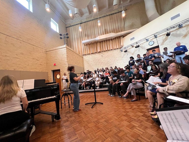 Mujer de pie dirigiendo a un grupo de músicos con partituras, piano en primer plano, entorno de sala beige.