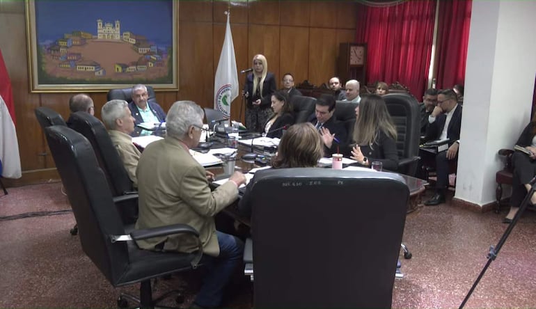 Hombre canoso con chaleco claro destaca en discusión, mientras mujer de negro gesticula, rodeados de asistentes en un ambiente formal.