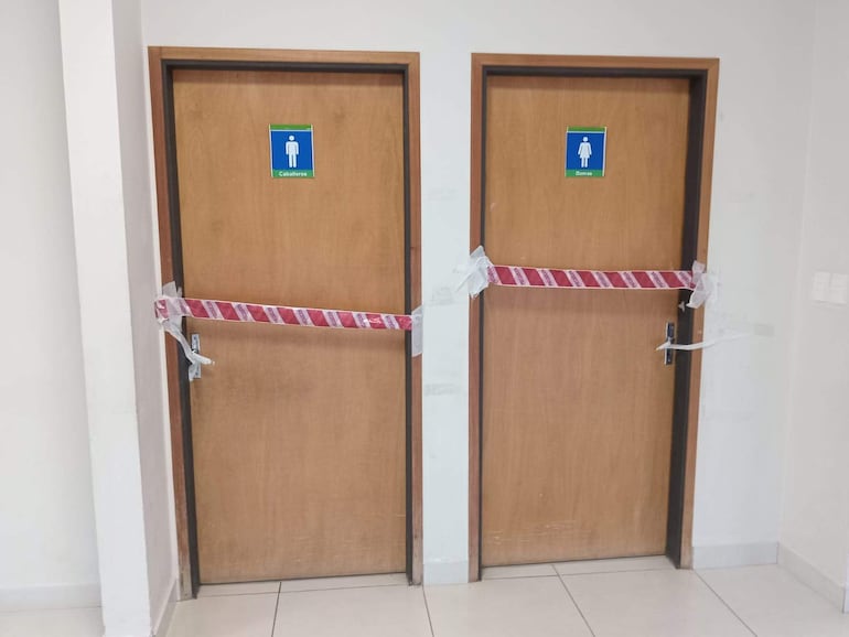 Puertas cerradas de baño en Clínica 12 de Junio, aseguradas con cinta de advertencia en un ambiente minimalista.