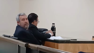 Hombre canoso en traje oscuro mira a la cámara, mientras que otro hombre con gafas se inclina hacia adelante en ambiente formal.