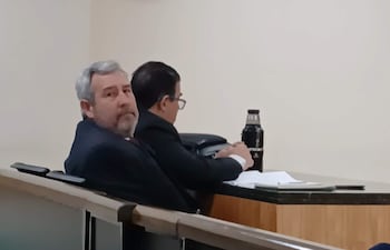 Hombre canoso en traje oscuro mira a la cámara, mientras que otro hombre con gafas se inclina hacia adelante en ambiente formal.