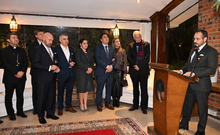 Vincenzo Turturro en saco oscuro habla a un grupo formal de hombres y mujeres, en un salón elegante con decoración sofisticada.