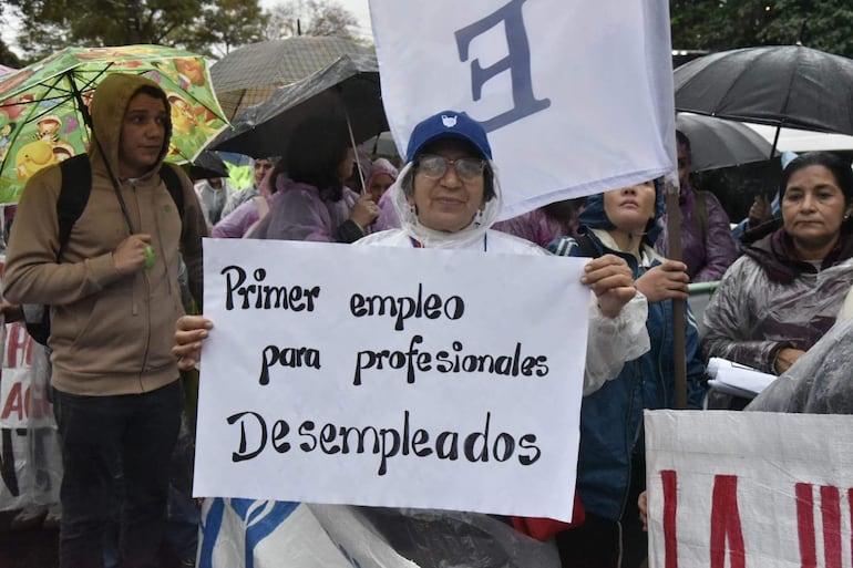 Mujer de cabello canoso con gafas y abrigo transparente sostiene un cartel sobre desempleo, rodeada de manifestantes bajo paraguas.
