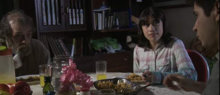 Héctor Silva con rostro tranquilo sobre una mesa con comida, acompañado de una niña y un joven en un comedor acogedor.