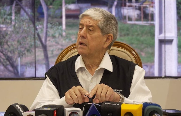 Carlos María Ljubetic, con chaleco oscuro y camisa blanca, gesticula en conferencia, rodeado de un entorno formal y vegetación visible.