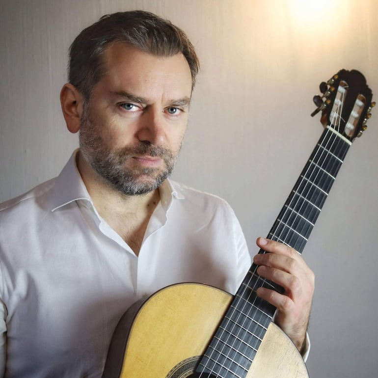 José Antonio Escobar con barba y cabellera corta, vestido con camisa blanca, sostiene una guitarra con expresión seria en un fondo neutro.