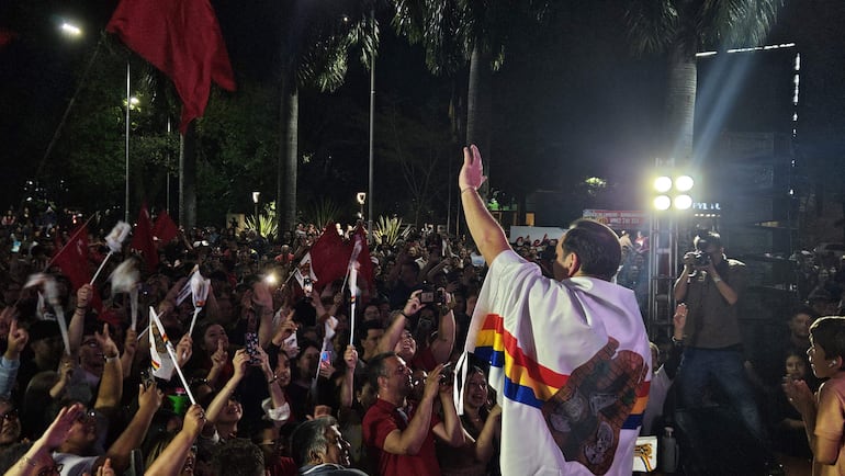 Sebastian Remesowski con camiseta blanca y abrigo colorido, saluda a una multitud emocionada con banderas rojas y luces brillantes.