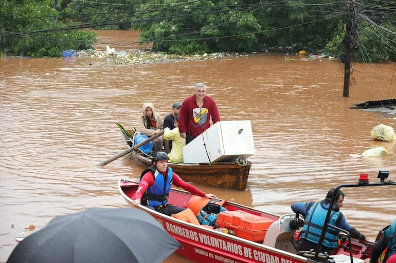 Hombre en bote de rescate con chaleco salvavidas, gente en canoa y electrodoméstico a la vista durante la inundación.