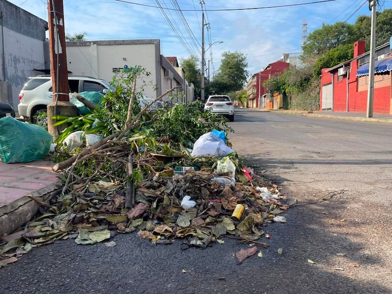 Calle con basura acumulada, un coche blanco estacionado y edificios de colores rojo y beige bajo un cielo despejado.