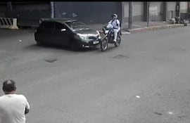 Hombre de espaldas en camiseta blanca observa un accidente entre un motociclista con uniforme y un automóvil negro.