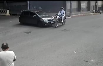 Hombre de espaldas en camiseta blanca observa un accidente entre un motociclista con uniforme y un automóvil negro.