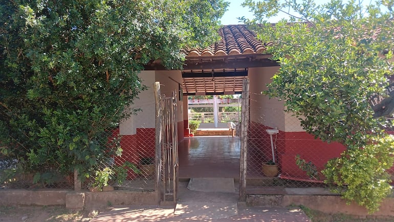 No hay personas visibles. Entrada de edificio blanco y rojo, con reja de metal y jardín verde al fondo.