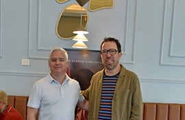 Martinessi en camiseta blanca y Peña Escobar con chaqueta verde, sonrientes en un ambiente moderno con sofá marrón claro.