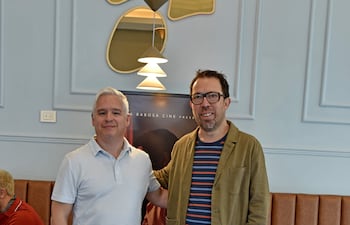 Martinessi en camiseta blanca y Peña Escobar con chaqueta verde, sonrientes en un ambiente moderno con sofá marrón claro.