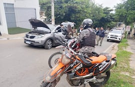 Policía Nacional en uniforme negro y chaleco, junto a SUV gris con capó abierto en calle con vegetación.