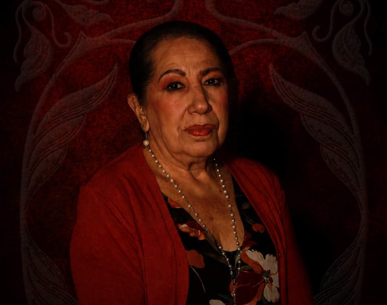 Teresa Barriocanal, mujer mayor con cabello oscuro y suéter rojo, posa seriamente frente a la cámara en un fondo rojo oscuro.
