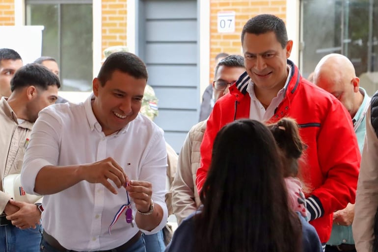 Diego Candia sonríe a una niña mientras sostiene un llavero, acompañado por Carlos Echeverría en chaqueta roja, en un ambiente festivo.