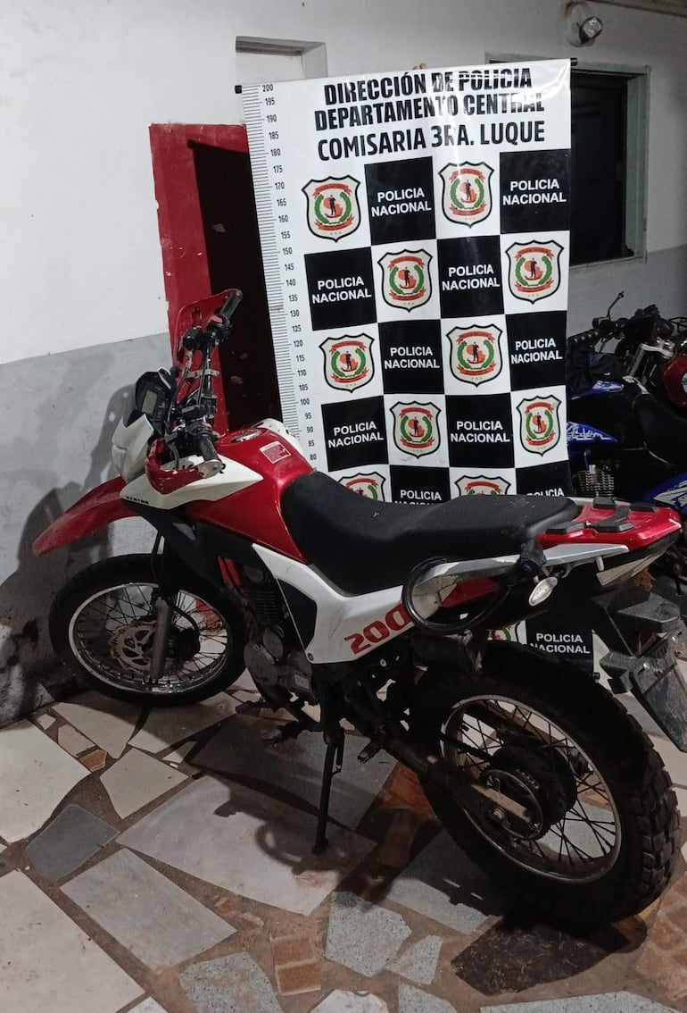 Motocicleta roja y blanca estacionada en una comisaría, con cartel de Policía Nacional en el fondo.