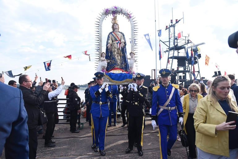 Cinco personas sostienen una imagen religiosa decorada mientras una multitud observa, con cielo parcialmente nublado de fondo.