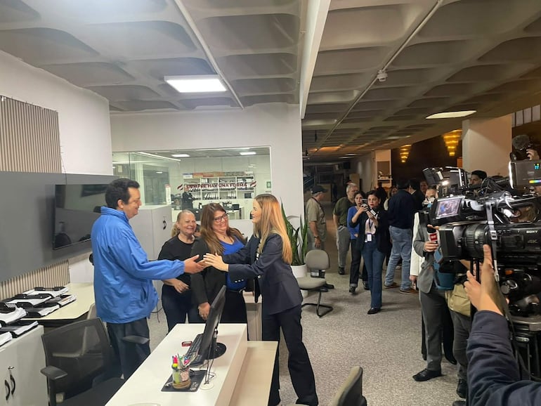 Soledad Núñez, con blazer oscuro, saluda a un hombre de chaqueta azul en un ambiente con otros observadores y cámaras.