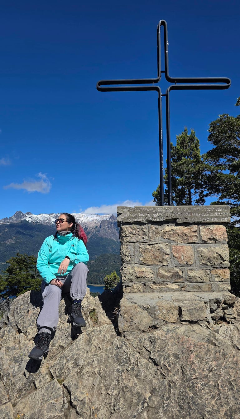 Mujer sentada sobre roca, con chaqueta azul y pantalones grises, frente al crucifijo en un paisaje montañoso con lago.
