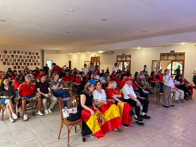 Grupo de personas con camisetas rojas y banderas de España, en un ambiente festivo y de expectación en la Casa de España.