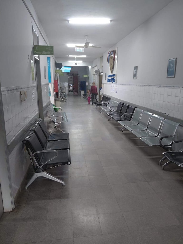 Mujer y niña caminan por un pasillo del hospital, mientras un hombre espera sentado en una silla de metal.