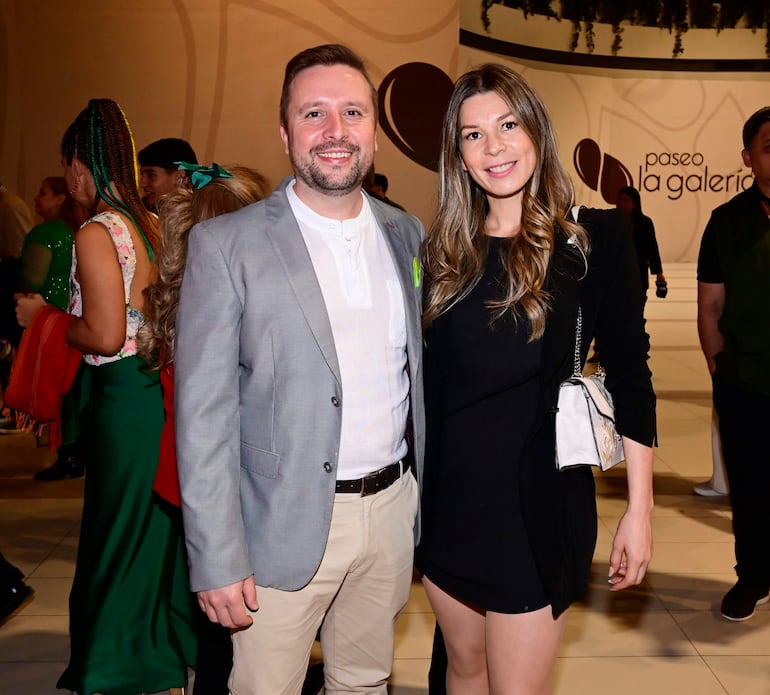 Hombre en saco gris y camiseta clara sonríe junto a mujer de cabello suelto, vestida de negro, en un evento festivo y colorido.