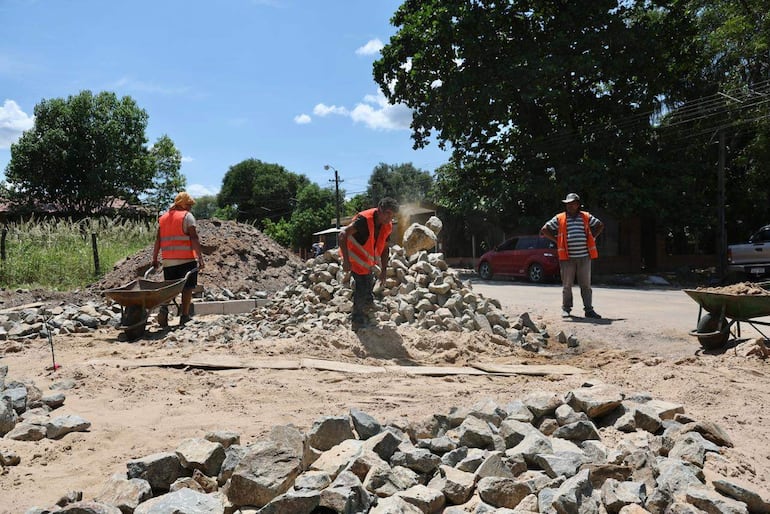 Dos trabajadores en chalecos naranjas, uno con pala y el otro con carretilla, observados por un tercero en un entorno de construcción.