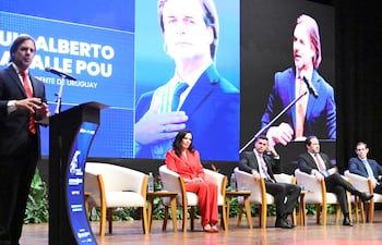 LU Alberto en traje negro y corbata roja, hablando frente a un micrófono. Una mujer con traje rojo escucha atentamente a su lado.