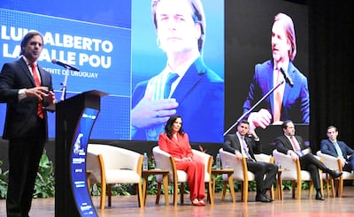 LU Alberto en traje negro y corbata roja, hablando frente a un micrófono. Una mujer con traje rojo escucha atentamente a su lado.