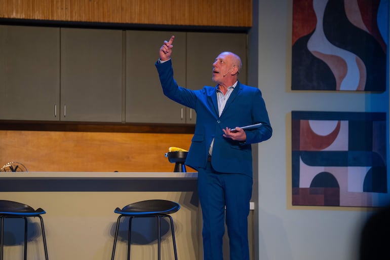 Hombre con traje azul sostiene una tablet mientras señala hacia el aire, con una cocina moderna como fondo.