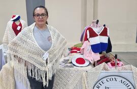 Lucia Jacquet, mujer de 50 años con gafas y chal claro, muestra prendas tejidas a mano en un ambiente de feria artesanal.