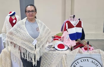 Lucia Jacquet, mujer de 50 años con gafas y chal claro, muestra prendas tejidas a mano en un ambiente de feria artesanal.