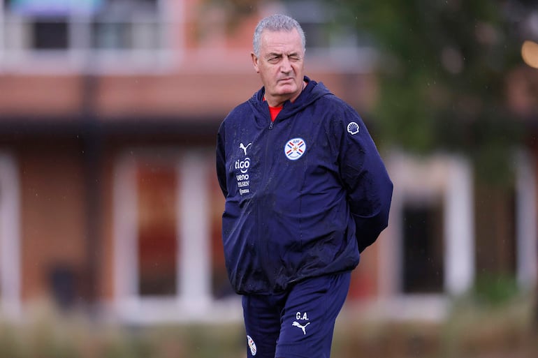 Gustavo Alfaro, vestido con abrigo institucional, observa serio el campo de entrenamiento bajo una leve lluvia.