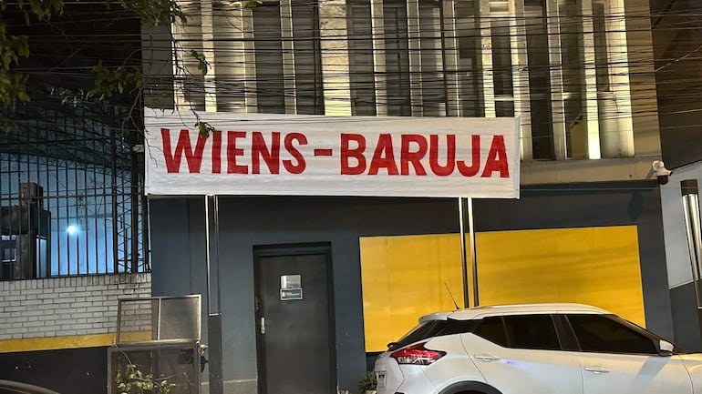 Letrero 'WIENS-BARUJA' en letras rojas sobre fondo blanco, frente a un edificio con puerta negra y pared amarilla.