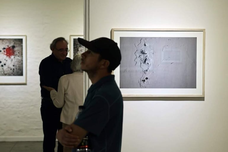 Hombre de perfil con gorra oscura escucha atentamente, mientras dos personas conversan al fondo en galería iluminada con obras contemporáneas.