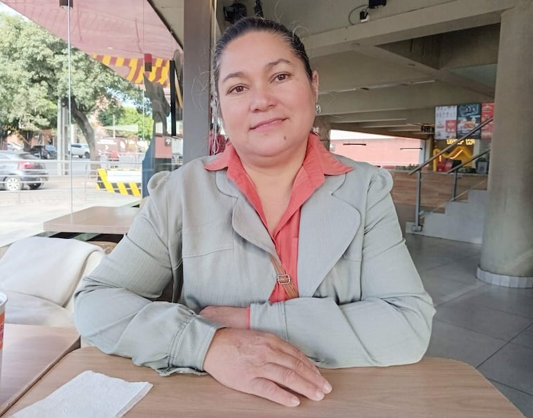 Clementina Ruiz Díaz Ojeda con chaqueta gris y camisa salmón sonríe en café, con manos cruzadas sobre la mesa en ambiente luminoso.
