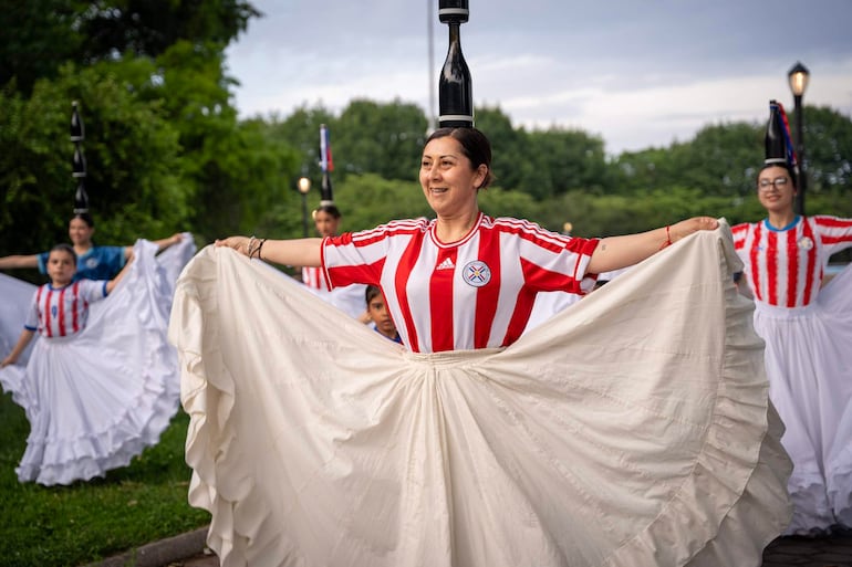 Mujeres paraguayas con camisetas rojas y blancas, sonriendo mientras sostienen faldas blancas en parque con faroles.