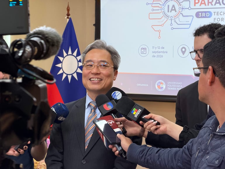 Hombre de mediana edad con cabello canoso y gafas sonríe mientras es entrevistado por periodistas, con bandera de Taiwán al fondo.