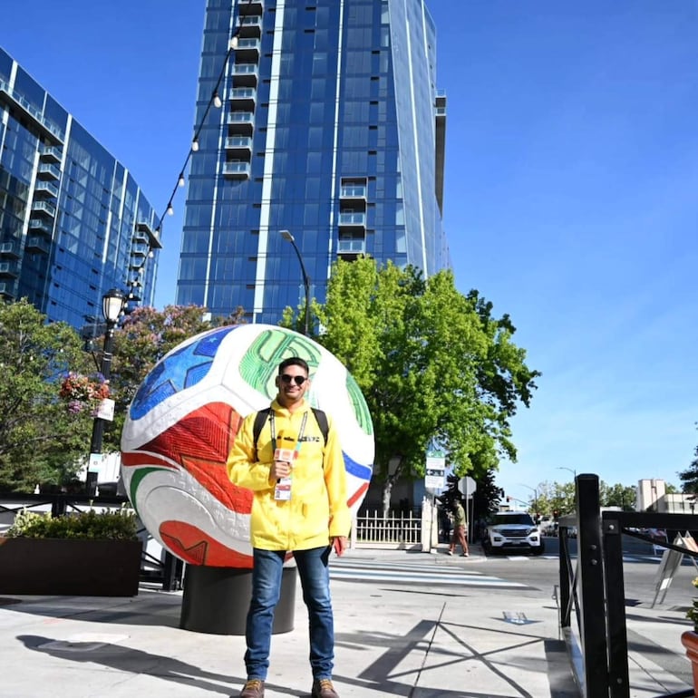 Hombre con abrigo amarillo y cámara sonríe frente a gran esfera decorativa de fútbol en un ambiente urbano moderno.